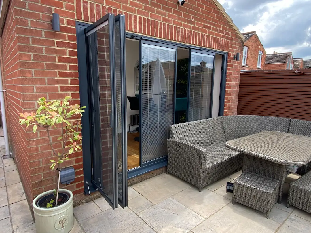 Partially open aluminium slim bifold doors (slide and pivot type) in black, with integral blinds, opening onto a patio